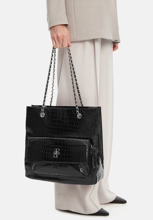 Osoba v béžovém obleku držící velkou černou kabelku s krokodýlím vzorem, stříbrnými řetízkovými popruhy a přední kapsou se závěsem z kovu.