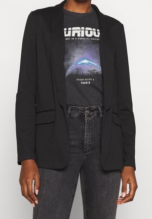 Person iført en sort blazer over en mørk grafisk t-shirt med et kosmisk design og mørke, højttaljede jeans.