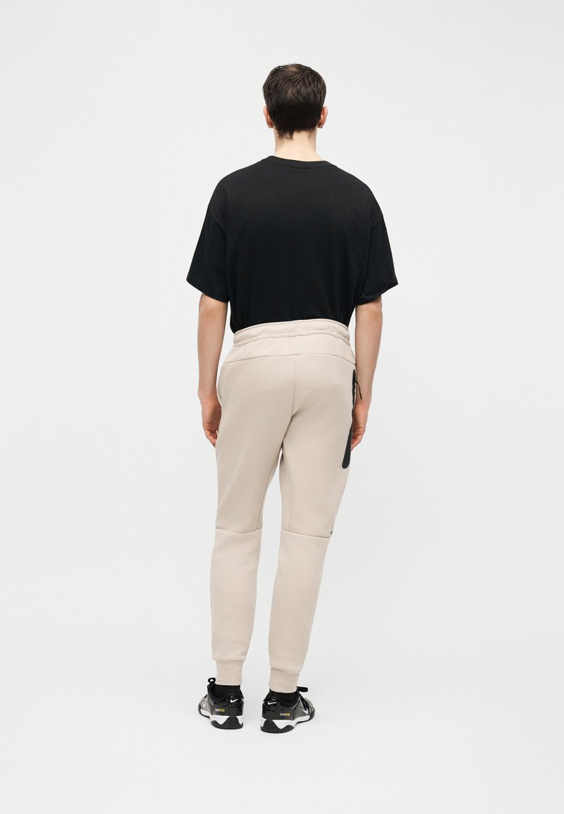 Hombre de pie de espaldas, vistiendo una camiseta negra, pantalones jogger beige y zapatillas negras sobre un fondo blanco liso.