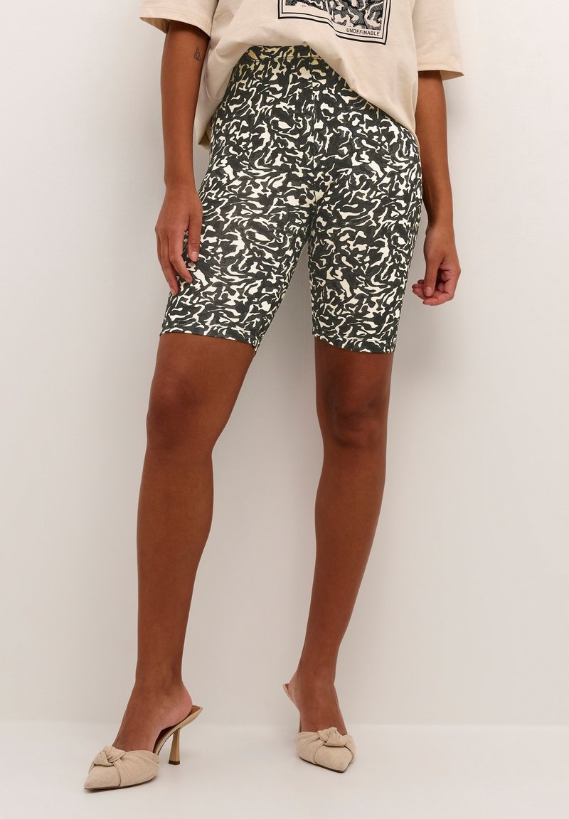 Personne portant un short cycliste taille haute noir et blanc à motif abstrait, un t-shirt graphique beige et des mules à talons beiges avec des nœuds.