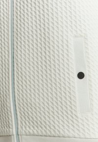 Gestreepte witte stof met een ruitpatroon, voorzien van een lichtblauwe rits en een zwarte knoopdetail op een zijzak.