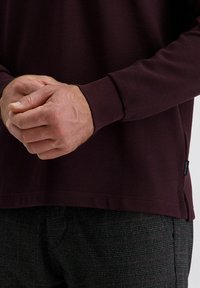 Mains serrées sur une chemise à manches longues rayée pourpre foncé et un pantalon à carreaux gris, montrant la texture du tissu et les détails des vêtements.