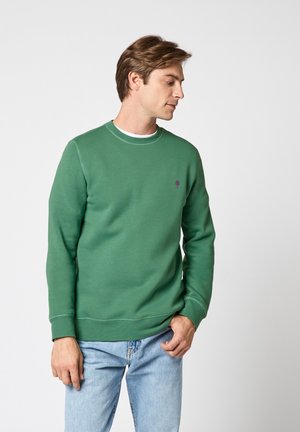 Homme portant un sweat-shirt vert à col rond et un jean bleu clair, regardant vers le bas et légèrement sur le côté sur un fond uni.