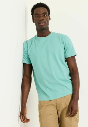 Junger Mann mit kurzen lockigen Haaren, der ein hellblaues T-Shirt und beige Hose trägt, an einer weißen Wand lehnt und nach vorne schaut.