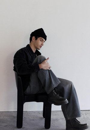 Fiatal férfi fekete sapkában, dzsekiben és bakancsban oldalra fordulva ül egy fekete széken, egyik térdét átölelve, egyszerű fehér fal előtt.