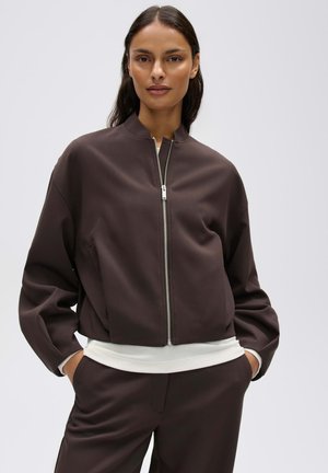 Donna con capelli lunghi e scuri che indossa una giacca marrone scuro con zip e pantaloni abbinati, con le mani nelle tasche su uno sfondo semplice.
