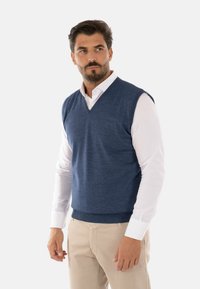 Gilet blu in misto lana con scollatura a V, indossato sopra una camicia bianca con colletto. Il gilet presenta un orlo a costine e una texture liscia.