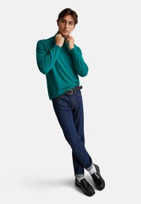 Turkusowy sweter z golfem, lekki materiał, zestawiony z ciemnoniebieskimi dżinsami. Czarny pasek i mokasyny dopełniają strój. Widoczne kontrastowe białe skarpetki.