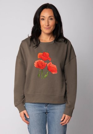 Vrouw met donker haar, gekleed in een bruine sweatshirt met drie felrode klaprozen, gecombineerd met blauwe jeans, staand tegen een effen achtergrond.