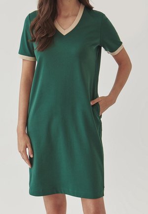 Femme portant une robe verte mi-longue avec une bordure beige au col en V et aux manches courtes, main dans la poche sur un fond uni.