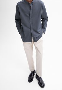 Camicia grigia a maniche lunghe con colletto alla coreana, abbinata a pantaloni beige chiaro e stivaletti neri. Stoffa liscia, linee pulite.