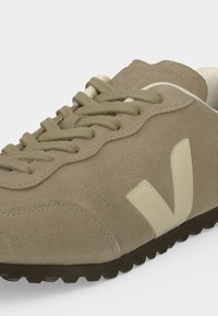 Baskets en daim beige avec lacets assortis, logo blanc "V" sur le côté et semelle noire rainurée pour l'adhérence et le style.