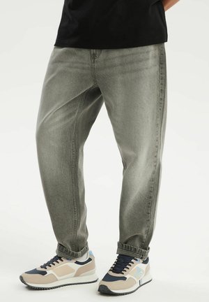 Dolna część ciała ubrana w szare, luźne jeansy oraz beżowe, czarne i białe trampki z jasnoniebieskim akcentem na białym tle.