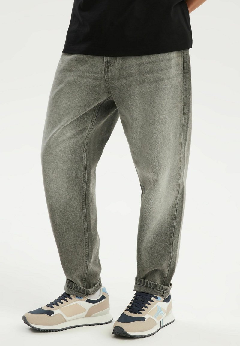 Dolna część ciała ubrana w szare, luźne jeansy oraz beżowe, czarne i białe trampki z jasnoniebieskim akcentem na białym tle.