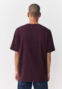 Maglietta di cotone color marrone con maniche corte, scollo rotondo e orlo dritto. Vista posteriore mostra un design pulito senza grafiche o motivi visibili.