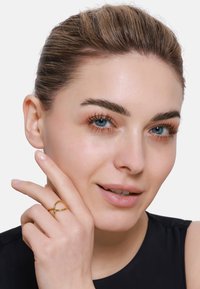 Goldring mit einem verflochtenen Design, der an einem Finger getragen wird. Das Model hat helle Haut und blaue Augen, mit glattem, zurückgebundenem Haar und neutralem Make-up.