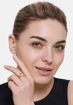 Goldring mit einem verflochtenen Design, der an einem Finger getragen wird. Das Model hat helle Haut und blaue Augen, mit glattem, zurückgebundenem Haar und neutralem Make-up.