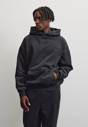 Homme aux cheveux tressés portant un sweat à capuche noir et un pantalon noir assorti, debout avec une main dans la poche sur un fond uni.