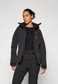 Giacca a vento nera con alto colletto, chiusura con zip e dettagli rossi; presenta una texture liscia e una silhouette aderente.