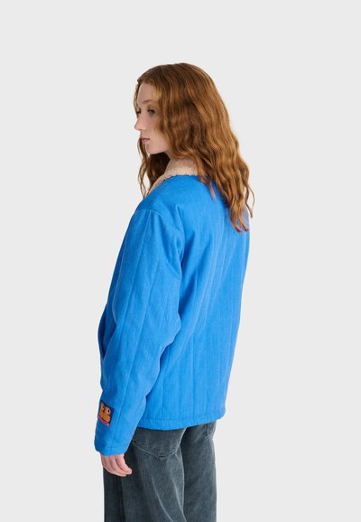 Femme aux longs cheveux roux portant une veste bleue avec un col en polaire et un pantalon gris, debout tournée vers la gauche sur un fond uni.