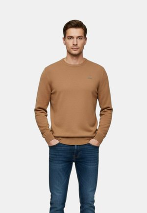 Uomo in piedi con le mani in tasca, che indossa un maglione marrone chiaro a maniche lunghe e jeans blu su uno sfondo chiaro uniforme.