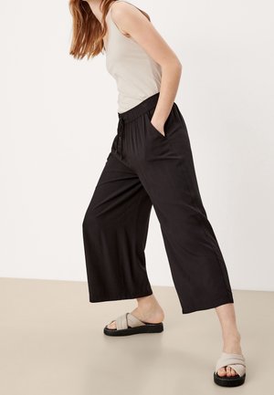 Pantalon large noir avec une taille élastique, des plis et des poches latérales. Associé à des sandales à enfiler gris clair avec une bride épaisse.