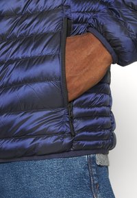 Marinblå puffjacka med kviltat mönster, sidoficka med dragkedja och elastiska ärmslut. Bärs över denimjeans med midjeband i grått.