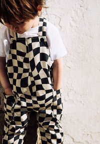 Salopette a quadri neri e crema con un motivo ondulato, caratterizzata da un design a taglio quadrato, spalline regolabili e tasche laterali.