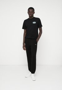 Črna majica s kratkimi rokavi z logotipom spredaj, v kombinaciji s črnimi trenirkami. Material se zdi mehak; model nosi bele superge. Enobarvne kombinacije skozi celotno obleko.