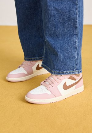 Pies con zapatillas Nike rosas, blancas y marrones con vaqueros azules, de pie sobre un suelo amarillo.