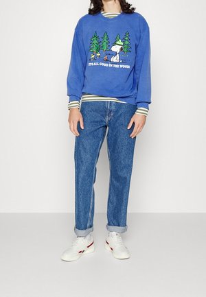 Personne portant un sweat bleu avec un motif Snoopy camping, un t-shirt rayé à manches longues en dessous, un jean bleu retroussé et des baskets blanches.