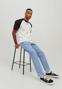 T-shirt z rękawami raglan w kolorze białym i czarnym, w połączeniu z jasnoniebieskimi dżinsami. Model siedzi na czarnym krześle barowym, nosząc czarne sneakersy.