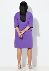 Fioletowa sukienka z krótkim rękawem o rozkloszowanym kroju, marszczona w talii, z długością do kolan. Noszona z jasnofioletowymi sandałami na wysokim obcasie.