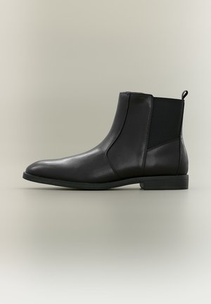 Bottine noire en cuir avec panneaux latéraux élastiques et languette de tirage, semelle plate et bout rond et lisse sur fond uni clair.