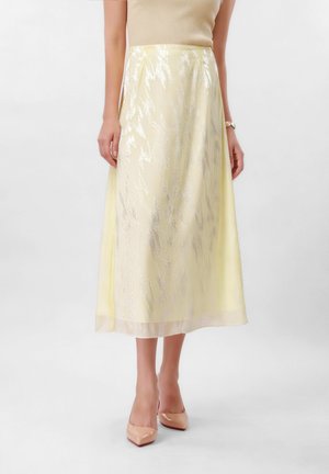 Femme portant une jupe midi jaune pâle avec un motif subtilement scintillant, un top beige sans manches, des escarpins pointus couleur nude et une montre-bracelet.