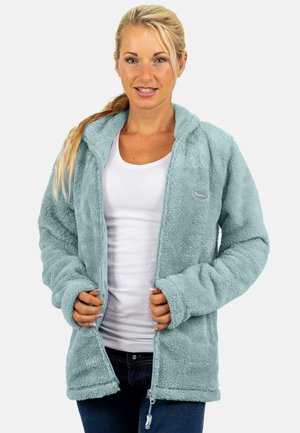 Blondine die een lichtblauwe fleecejas draagt over een wit shirt en donkere spijkerbroek, met beide handen de rits van de jas vasthoudend.
