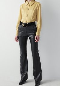 Gul langærmet skjorte med knaplukning, parret med høj talje sorte faux læder flared bukser og et bredt sort bælte med en gylden spænde.