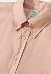 Chemise boutonnée rose clair en coton, dotée d'un col pointu, de détails cousus et de quatre boutons de fermeture. Logo brodé à l'avant.