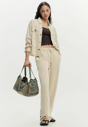 Young woman in beige jacket and pants, black crop top, black sandals, holding large greenish handbag with colorful charms.