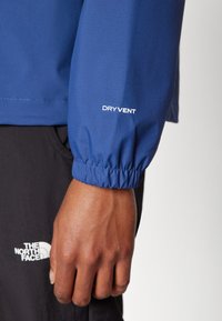 Veste imperméable bleue avec poignets élastiques et étiquette "DRY VENT". Comprend un tissu texturé et un design à plusieurs couches avec un logo visible au niveau du poignet.