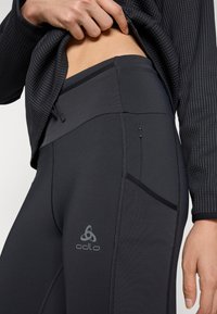 Juodos sportinės legging'os su lygaus paviršiaus tekstūra, su užtrauktuku užsegama kišenė, aukšta juosmens linija ir Odlo logotipu kairėje šlaunies pusėje.