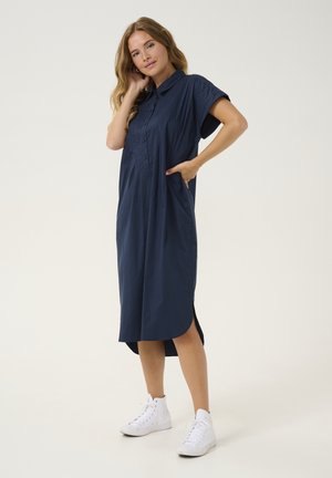 Marineblaue Kurzarm-Kleidbluse mit Knopfleiste, Seitentaschen und leicht gebogtem Saum. Tragen mit weißen Sneakers.