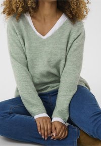Jasnozielony sweter z prążkowanej dzianiny z dekoltem w serek, zakładany na białą koszulę. Modelka nosi niebieskie dżinsy i siedzi z dłońmi złożonymi na udach.