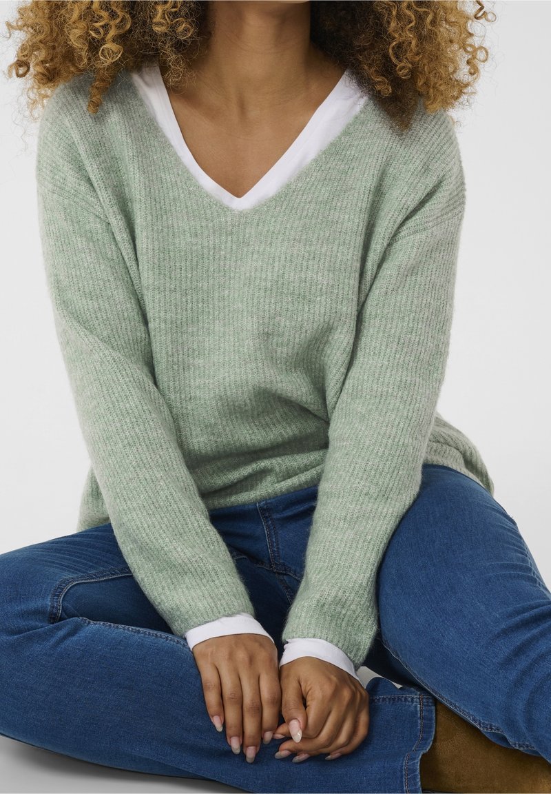 Jasnozielony sweter z prążkowanej dzianiny z dekoltem w serek, zakładany na białą koszulę. Modelka nosi niebieskie dżinsy i siedzi z dłońmi złożonymi na udach.