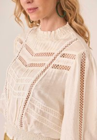 Blusa blanca de tela texturizada, con un cuello alto y un intrincado diseño de entramado. Las mangas largas y los acentos en forma de concha añaden dimensión.