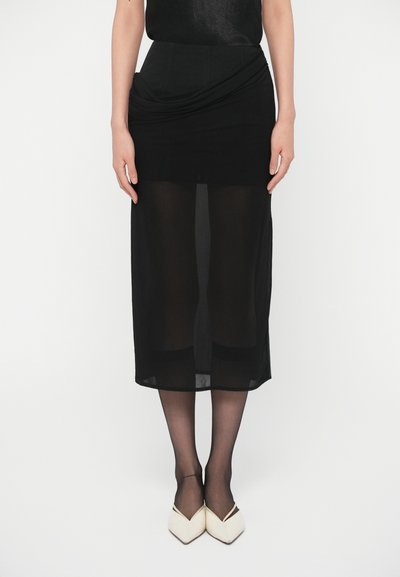 Jupe midi noire avec une superposition transparente. Présente un design drapé à la taille et une sous-couche ajustée. Associée à des chaussures blanches à bout pointu.