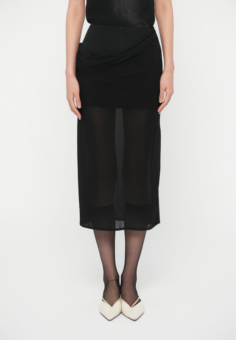 Saia midi preta com uma sobreposição translúcida. Apresenta um design drapeado na cintura e uma camada inferior ajustada. Combinada com sapatos brancos de bico fino.