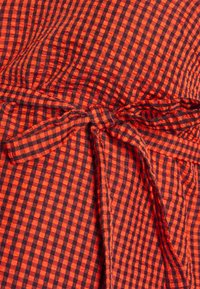 Tecido em padrão quadriculado vermelho e preto com detalhe na cintura amarrado. A textura parece leve com um padrão tecido visível.