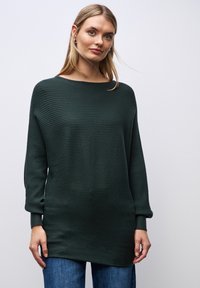 Dunkelgrüner, gerippter Pullover mit weitem Ausschnitt und langen Ärmeln, aus einem weichen Material gefertigt, kombiniert mit blauen Jeans.