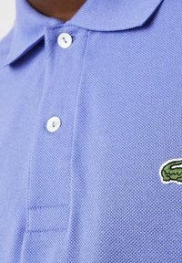 Polotröja i ljuslila med en texturerad tyg och tre vita knappar. Har en grön krokodilmärke på vänster bröstområde.